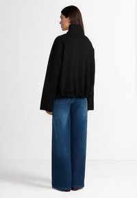 Črna majica z visokim ovratnikom, sproščenega kroja in nabranim spodnjim robom, kombinirana z modrimi širokimi kavbojkami. Gladek tekstura, minimalen dizajn.