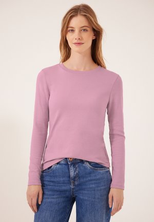 Femme aux cheveux châtain clair portant un t-shirt rose à manches longues légèrement rentré dans un jean bleu, debout devant un fond uni.