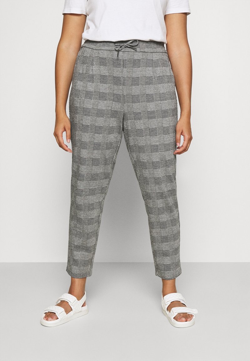 Personne portant un pantalon gris à carreaux longueur cheville avec une taille à cordon, un t-shirt blanc et des sandales blanches, debout devant un fond uni.