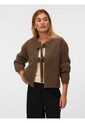 Femme portant un cardigan marron en maille avec deux liens à l'avant sur un top crème et un pantalon noir, debout avec une main sur la hanche.