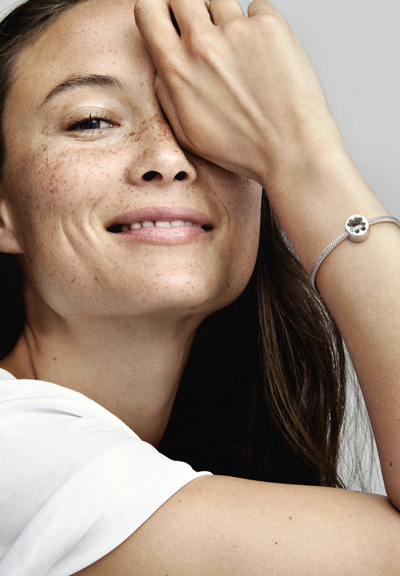 Bracelet en argent avec un charme rond en forme de fleur. Le modèle a de longs cheveux, porte une chemise blanche et met en valeur le bracelet à son poignet.