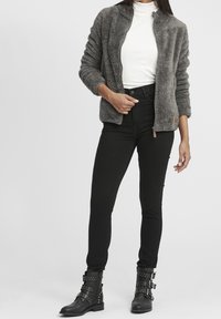 Harmaa pörröinen vetoketjullinen takki valkoisen poolopaidan päällä, yhdistettynä korkeavyötäröisiin mustiin farkkuihin ja mustiin niitattuihin nilkkabootseihin.