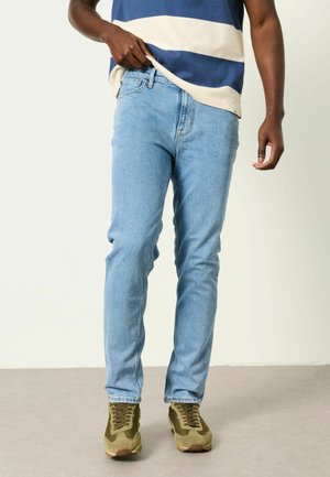 Osoba ubrana w jasnoniebieskie dżinsy, oliwkowozielone trampki i niebiesko-białą paskowaną koszulę, stojąca na szarej podłodze.