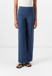 Pantalons larges en tissu bleu foncé avec une texture lisse. Design taille haute sans matériel visible. Associés à des sandales noires.
