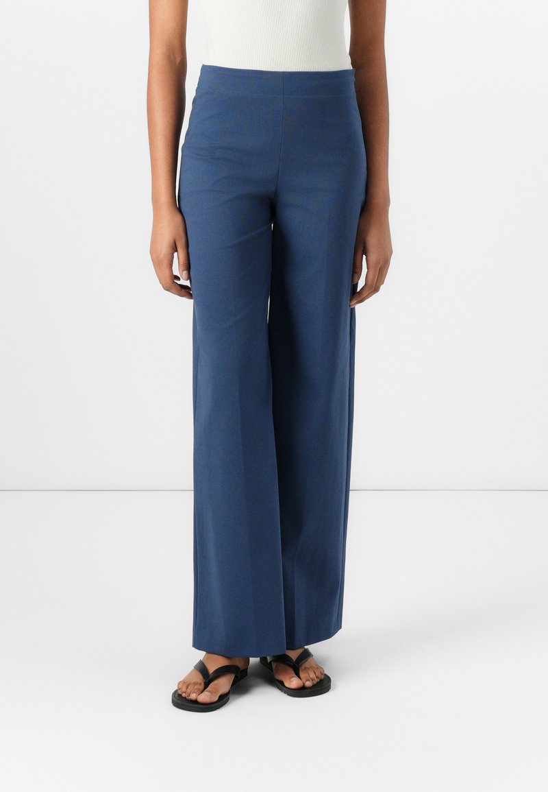 Pantalons larges en tissu bleu foncé avec une texture lisse. Design taille haute sans matériel visible. Associés à des sandales noires.