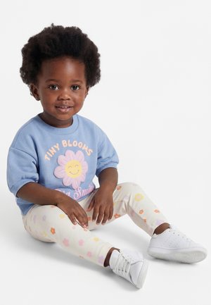 Maluch z kręconymi włosami ubrany w niebieską koszulkę "Tiny Blooms", kwieciste legginsy i białe trampki, siedzący na białej podłodze i uśmiechający się.