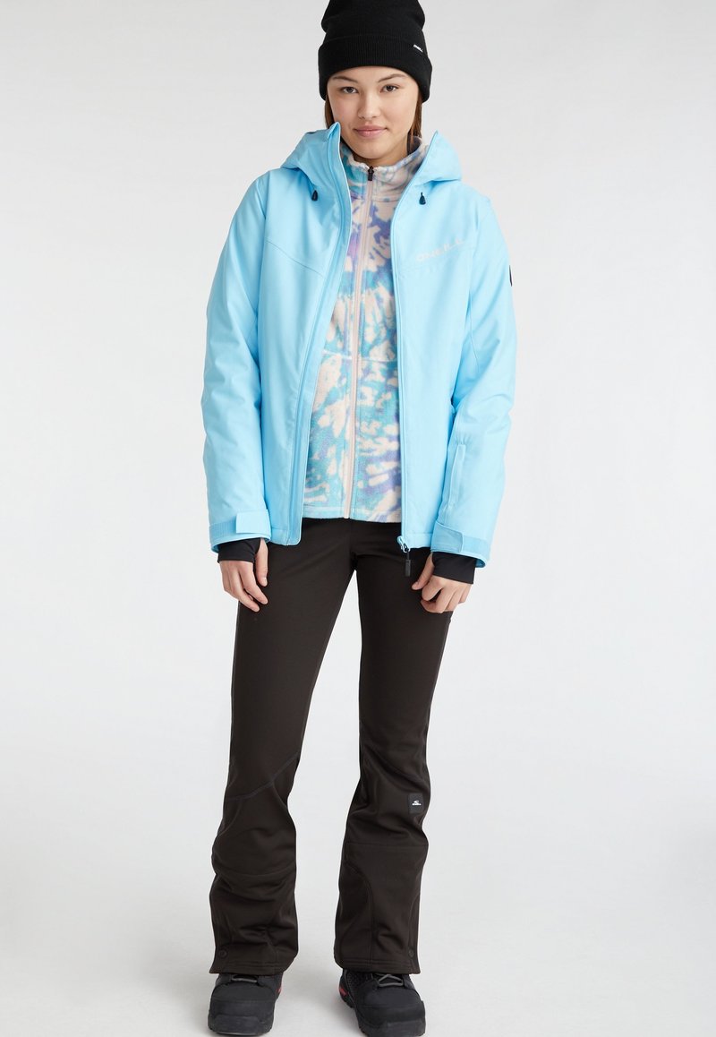 Giacca isolante blu cielo con zip frontale, cappuccio e polsini neri a contrasto, indossata sopra un pile a fantasia, abbinata a pantaloni neri e stivali.