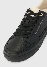 Musta tennari, jossa on nahkainen päällinen, pyöreä varvas ja tasainen kumipohja. Varusteena mustat nauhat ja kultainen logo sivulla. Pintatextuuri on sileä ja matta.