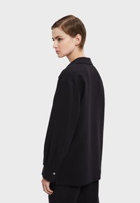 Musta pitkähihaine paita, jossa on kaulus, rento leikkaus, nappikiinnitys hihansuissa ja sileä pinta. Kangas näyttää pehmeältä ja laskeutuu kauniisti.