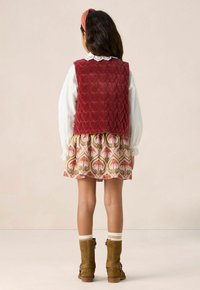 Gilet matelassé rouge avec motif de cœurs, blouse blanche avec col en dentelle, jupe à motifs, bottines marron, et chaussettes crémeuses avec bandes colorées.