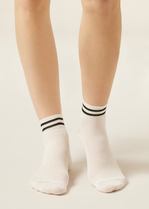 Jambes portant des chaussettes blanches aux chevilles avec deux rayures noires près du haut, debout sur un fond clair et neutre.