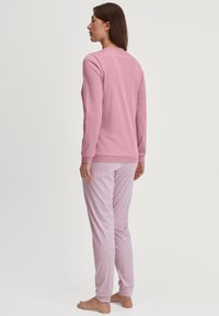 Vrouw die blootsvoets met haar gezicht weg staat, gekleed in een lange mouwen roze top en lichtroze gestreepte pyjamabroek tegen een effen achtergrond.