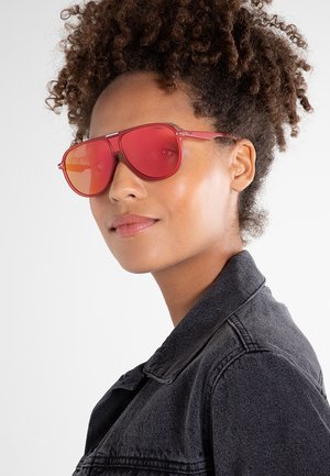 Giovane donna con capelli ricci indossa occhiali da sole aviator rossi e una giacca di jeans nera, guardando leggermente di lato con un'espressione neutra.