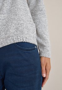 Grå strukturerad tröja med ett vitt mönster, parat med mörkblå denimjeans med en framficka och metallknapp som accent.