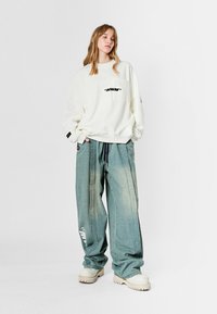 Sudadera blanca de gran tamaño con un bolsillo en el pecho y texto negro, combinada con pantalones de mezclilla sueltos de color azul claro con acentos laterales. Calzado chunky.