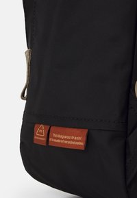 Zaino nero realizzato in nylon riciclato. Presenta una superficie testurizzata, un'impugnatura e due etichette rosse che indicano il marchio e le informazioni sul materiale.