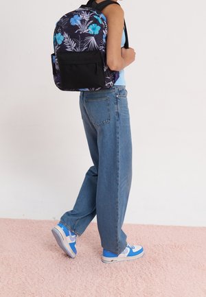 Zaino floreale nero con ibisco blu e foglie di palma bianche, dotato di tasca anteriore e spallacci regolabili, abbinato a jeans blu larghi e sneakers.