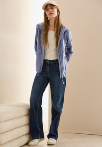 Veste zippée bleu clair, chemise blanche, jean large bleu marine, casquette beige et baskets blanches. Textures douces avec des éléments de design fonctionnels.