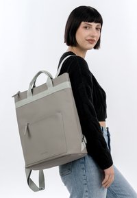 Junge Frau mit kurzen dunklen Haaren, die einen großen grauen Rucksack über einer Schulter trägt, bekleidet mit einem schwarzen Pullover und blauen Jeans.