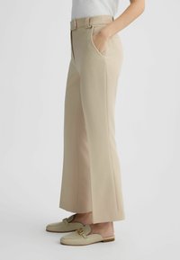 Pantaloni beige a gamba larga con design sartoriale, tasche laterali e cintura. Tessuto morbido con linee pulite, abbinati a scarpe slip-on di colore chiaro.