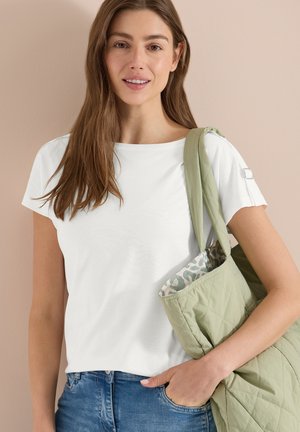 Jeune femme en haut blanc à manches courtes et jeans bleus porte un sac cabas matelassé vert clair avec une doublure à motifs sur l'épaule.