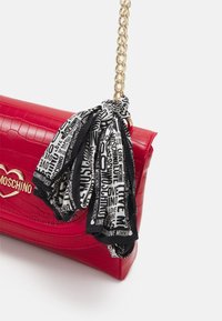 Borsa in pelle sintetica rossa effetto coccodrillo con tracolla in catena dorata, decorata con una sciarpa stampata in bianco e nero legata al manico.