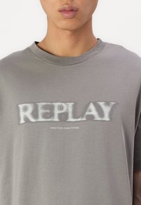 Grå bomull T-skjorte med korte ermer, med en stor hvit "REPLAY" logo og mindre "NINETEEN EIGHTYONE" tekst under. Glatt tekstur.