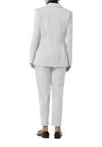 Traje gris claro con blazer ajustado y pantalones de corte slim. La tela parece suave con una textura sutil. Unos zapatos negros brillantes completan el look.
