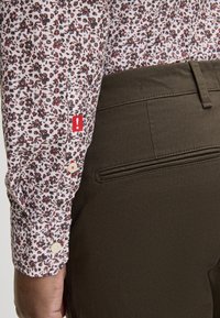 Langarmshirt met een witte basis en een bloemenpatroon in rood en marineblauw. Bruine broek met een achterzak en textuurstof. Rode accenten op de manchet.