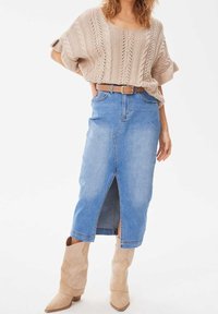 Suéter beige de punto con diseño de trenzado, combinado con una falda midi de mezclilla azul clara con una abertura frontal. Cinturón tostado y botas de vaquero tostadas.