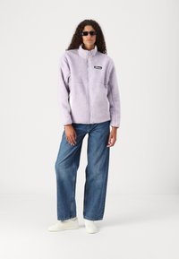 Lilla fleecejakke med høy krage, glidelås foran og svart lapp. Kombinert med løse blå jeans og hvite joggesko.