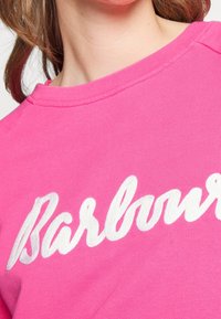 Sweatshirt rose avec le texte "Barbour" brodé en blanc. Le tissu est lisse, avec un col rond et des détails de couture subtils.