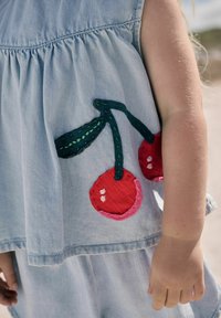 Haut en denim bleu clair avec un design froncé, orné de cerises rouges brodées et de feuilles vertes sur le côté. Texture douce.