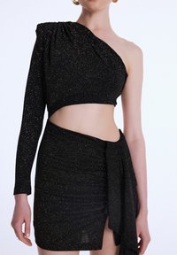 Černý třpytivý crop top s jedním dlouhým rukávem, asymetrickým výstřihem a nabíranou sukní s drapovaným akcentem. Hladká textura, přiléhavý design.