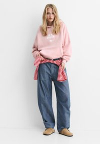 Femme blonde portant un sweat à capuche rose surdimensionné avec du texte, une chemise à rayures larges nouée à la taille, un jean bleu ample et des chaussures slip-on beiges, debout.