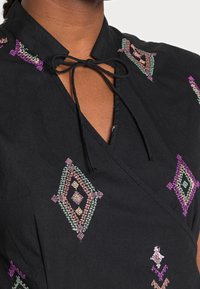 Abito nero con motivi a diamante colorati ricamati e un fiocco legato al colletto su pelle scura.