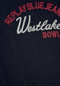 Mörkblått tyg med broderad röd och vit text: "REPLAY BLUE JEANS WESTLAKE BOWL." Texten är styliserad i olika typsnitt och storlekar.