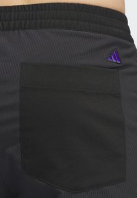 Artimiausias juodų sportinių šortų vaizdas su dideliu užpakaliniu kišeniu ir maža violetine Adidas logo arti juosmens.