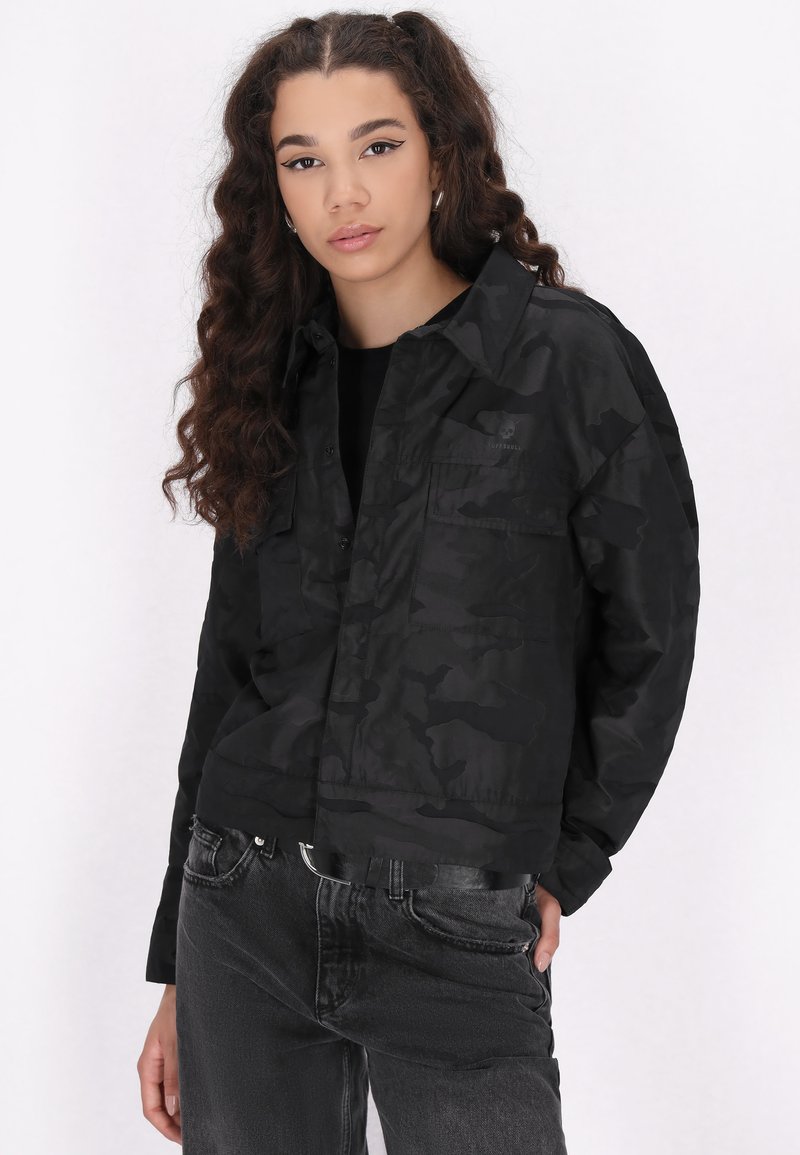 Zwarte camouflage jas met een kraag, twee voorzakken en een iets kortere pasvorm. Draagt met donkergrijze jeans. Gladde textuur.