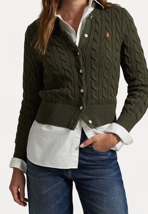 Femme portant un cardigan en maille torsadée vert olive sur une chemise blanche à boutons et un jean bleu, main dans la poche, debout devant un fond uni.