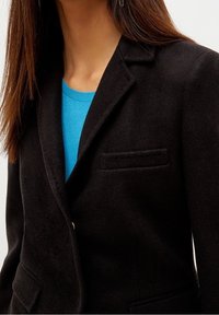 Svart ullblazer med utvändig krage, en enkel ficka och en knapp. Bärs över en ljusblå, texturerad tröja.