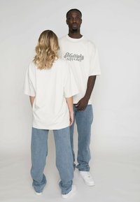 Přepychové bílé trička s nápisem "Multiply Apparel", spárované s modrými džíny. Modelové stojící zády k sobě na bílém pozadí.