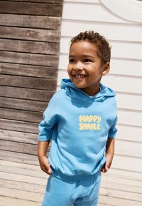 Svetle modrá mikina s krátkým rukávem, se nápisem "HAPPY SMILE" v žlutém písmu. Odpovídající šortky, oboje vyrobené z měkké látky.