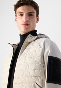 Light beige quilted jacket with a hood, featuring a black zippered pocket on the left arm and a smooth black collar.