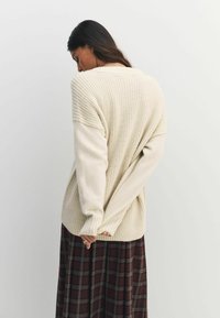 Kreemne kudumisega sviiter, millel on soonikkoes ja pikad varrukad, kombineerituna tumeda ruudulise seeliku ja tagantvaatega.