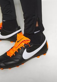 Černé kožené fotbalové kopačky s bílým logem Nike, oranžovými detaily a jasně oranžovými tkaničkami. Černé kalhoty se zipy na kotnících.