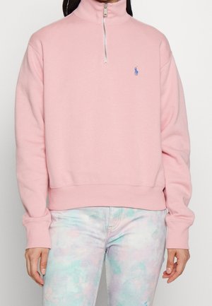Sweatshirt rose à fermeture éclair avec un col haut, des poignets et un ourlet côtelés. Petit logo sur la poitrine gauche. Porté avec un pantalon en tie-dye pastel.