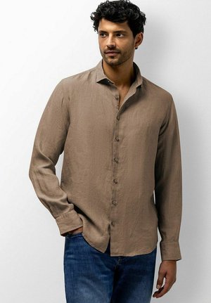 Mann mit lockigem Haar, der ein langärmeliges braunes Hemd mit Knöpfen und blaue Jeans trägt und mit einer Hand in der Hosentasche an einer weißen Wand steht.