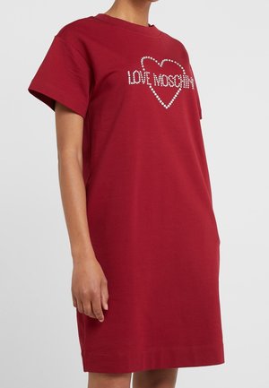 Kobieta w czerwonej sukience z krótkim rękawem z kryształowym sercem i napisem "LOVE MOSCHINO" na piersi.