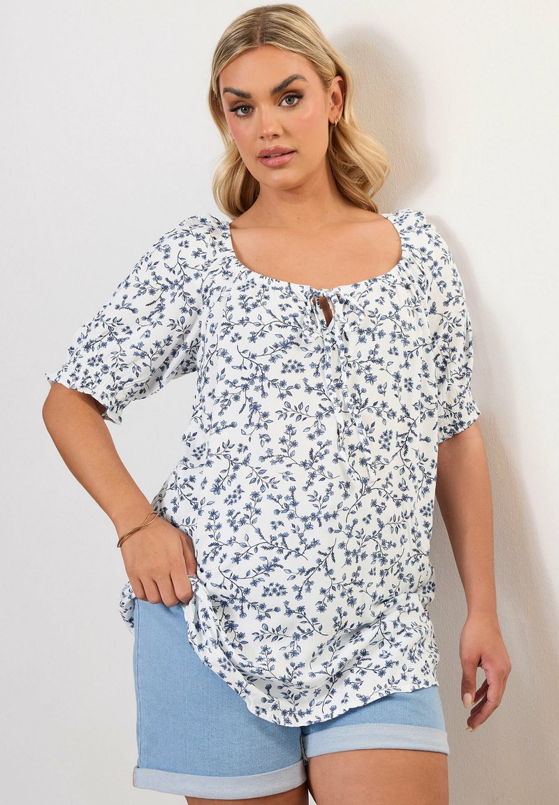 Blusa floreale bianca con motivi blu, maniche corte a palloncino e scollatura quadrata. Abbinata a pantaloni shorts in denim blu chiaro.